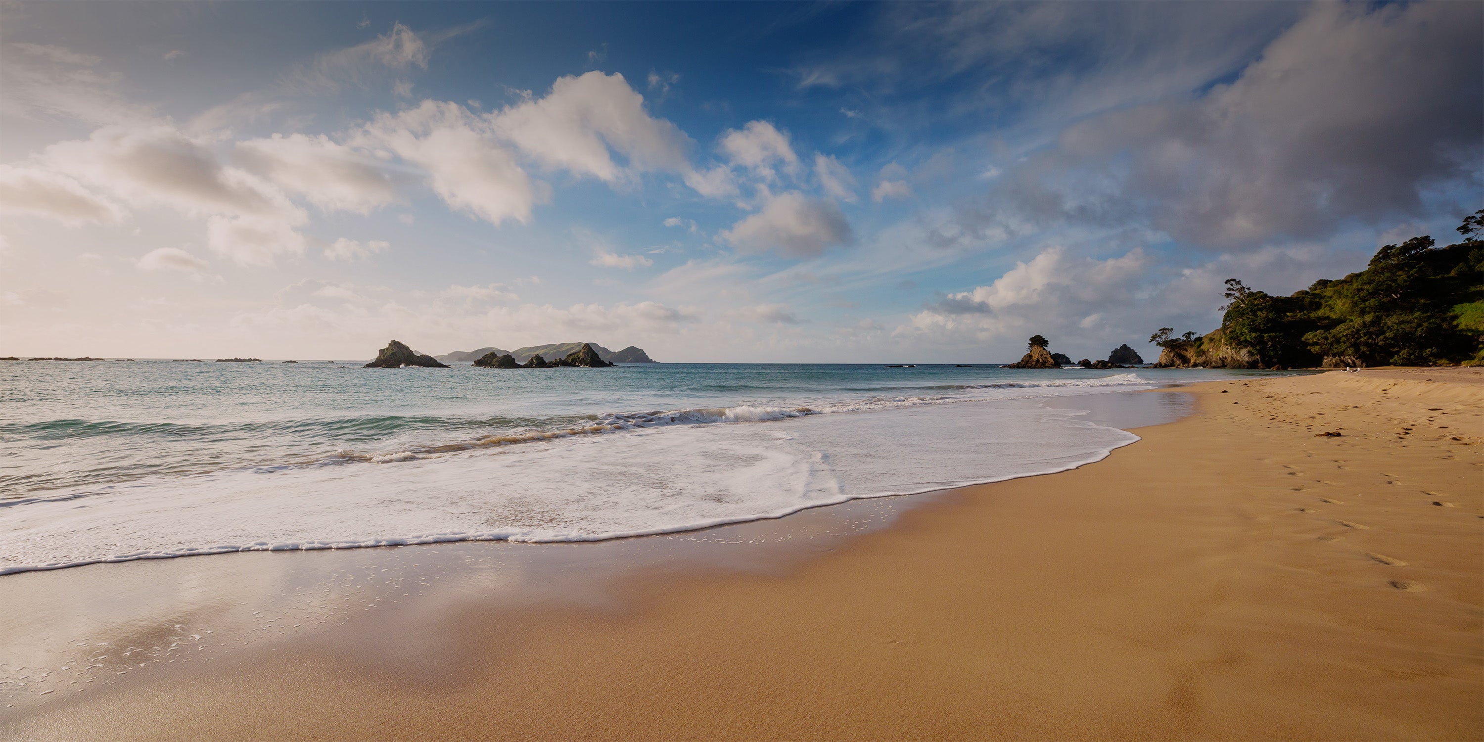 Beach scene with waves crashing onto the shore under a blue sky with clouds.
