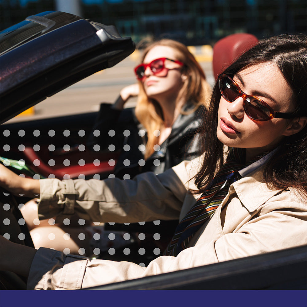 Two women in a convertible car, one driving and the other sitting in the passenger seat, wearing sunglasses.
