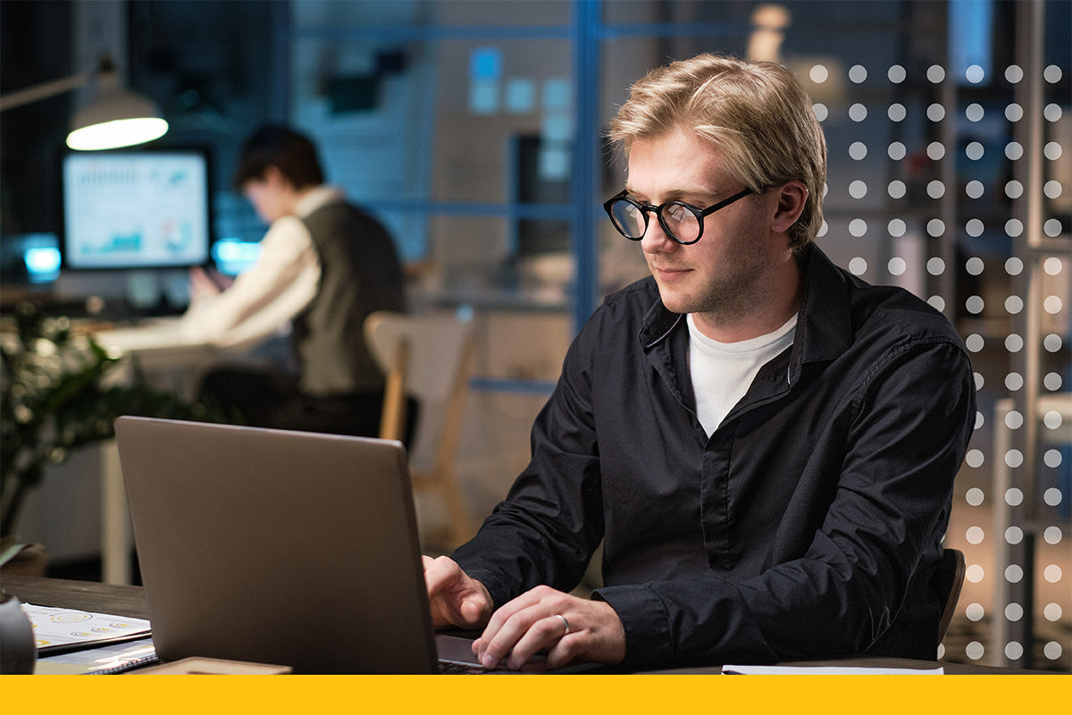 Man working on a laptop in an office setting with another person in the background.